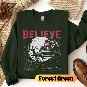 May include: A forest green sweatshirt with a graphic of Santa Claus flying in a UFO with reindeer. The text "BELIEVE" is printed in red above the graphic. The sweatshirt is on a white surface.