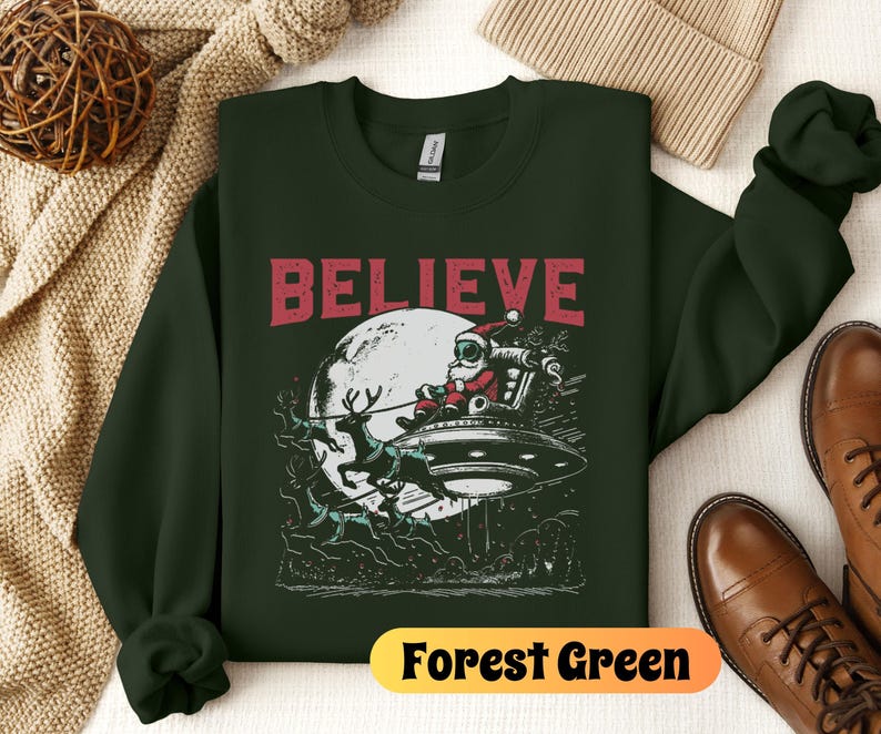 May include: A forest green sweatshirt with a graphic of Santa Claus flying in a UFO with reindeer. The text "BELIEVE" is printed in red above the graphic. The sweatshirt is on a white surface.