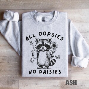 May include: Light grey sweatshirt with a raccoon graphic holding a drink and a sunflower. The text "ALL OOPSIES NO DAISIES" is printed on the front. The sleeves are cuffed. The word "ASH" is printed at the bottom.