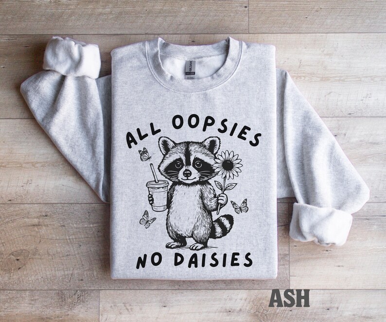 May include: Light grey sweatshirt with a raccoon graphic holding a drink and a sunflower. The text "ALL OOPSIES NO DAISIES" is printed on the front. The sleeves are cuffed. The word "ASH" is printed at the bottom.