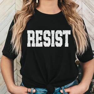 May include: Black t-shirt featuring the word "RESIST" in large, distressed white lettering. The shirt has a crew neck and short sleeves. The model is wearing blue jeans.