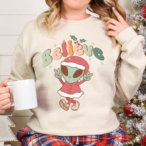 May include: A cream-colored sweatshirt with a festive alien wearing a Santa hat and the word "Believe" in colorful letters. The alien is holding up two fingers in a peace sign.
