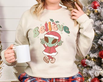 Puede incluir: Una sudadera de color crema con un extraterrestre festivo que lleva un gorro de Papá Noel y la palabra "Believe" en letras de colores. El extraterrestre levanta dos dedos en señal de paz.