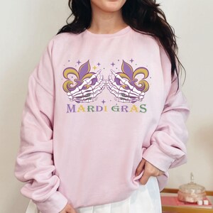 May include: Pink sweatshirt with a Mardi Gras design featuring two skeletal hands holding fleur-de-lis. The text "Mardi Gras" is printed below the design.