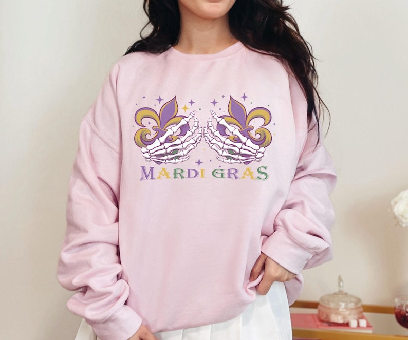 May include: Pink sweatshirt with a Mardi Gras design featuring two skeletal hands holding fleur-de-lis. The text "Mardi Gras" is printed below the design.