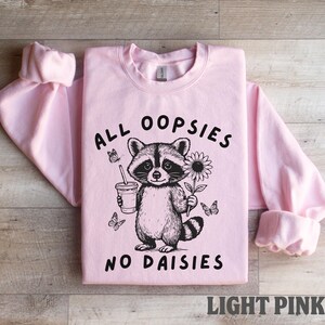May include: Light pink sweatshirt with the text "ALL OOPSIES NO DAISIES" and a raccoon graphic holding a drink and a flower. The graphic is black and white. The sweatshirt is displayed on a wooden surface.