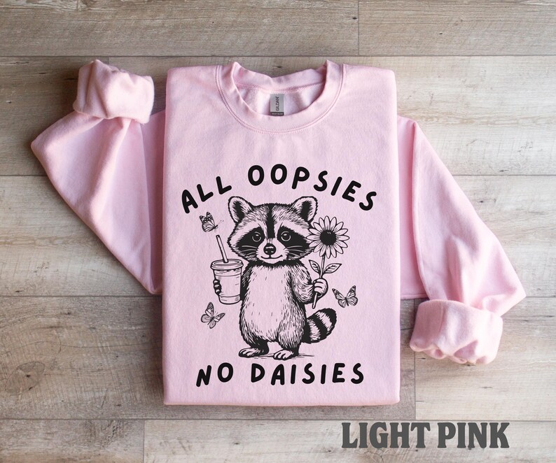 May include: Light pink sweatshirt with the text "ALL OOPSIES NO DAISIES" and a raccoon graphic holding a drink and a flower. The graphic is black and white. The sweatshirt is displayed on a wooden surface.
