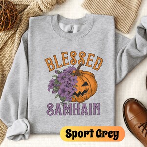 以下が含まれることがあります： グレーのトレーナーに、紫とオレンジの文字で「Blessed Samhain」と書かれています。トレーナーの中央には、紫の花がついた、アニメーションのジャック・オ・ランタンが描かれています。トレーナーには「Sport Grey」のラベルが付いています。