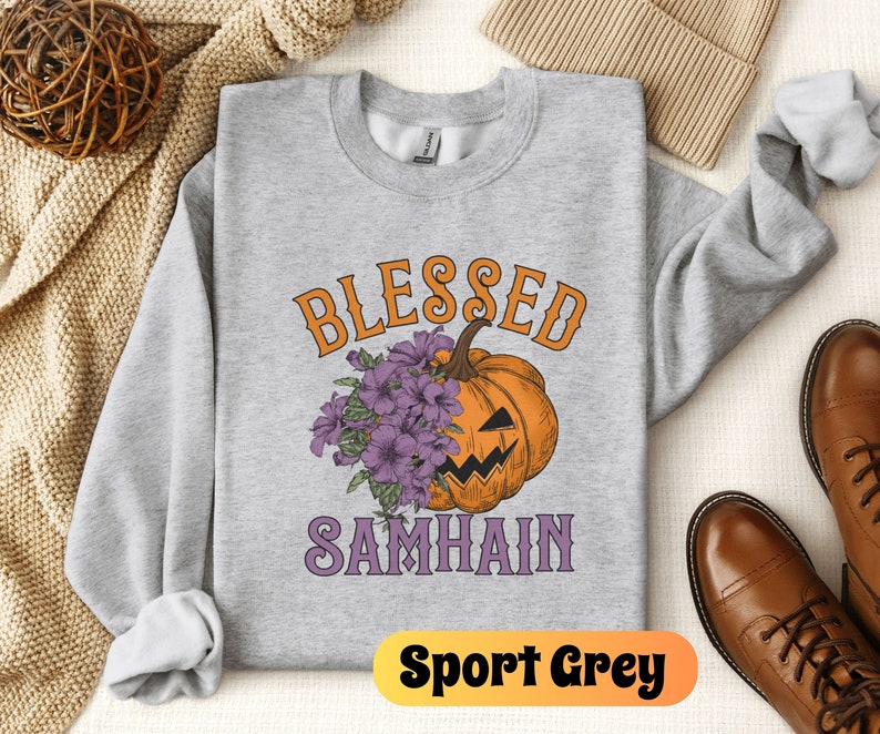 以下が含まれることがあります： グレーのトレーナーに、紫とオレンジの文字で「Blessed Samhain」と書かれています。トレーナーの中央には、紫の花がついた、アニメーションのジャック・オ・ランタンが描かれています。トレーナーには「Sport Grey」のラベルが付いています。