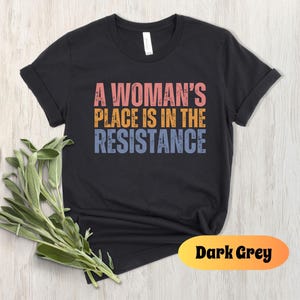 May include: Dark grey t-shirt with the text "A Woman's Place Is In The Resistance" in pink, orange, and blue. The shirt has rolled-up sleeves and a crew neck. A sprig of green leaves is in the lower left corner.