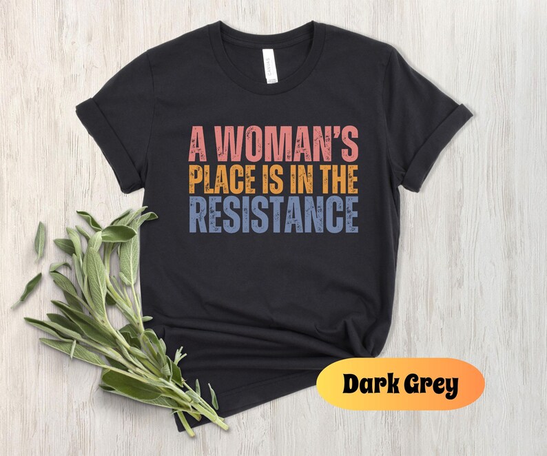 May include: Dark grey t-shirt with the text "A Woman's Place Is In The Resistance" in pink, orange, and blue. The shirt has rolled-up sleeves and a crew neck. A sprig of green leaves is in the lower left corner.