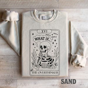 Puede incluir: Sudadera color arena con un dise&ntilde;o de carta de tarot en blanco y negro. La carta representa un esqueleto sosteniendo una taza, con el texto "WHAT IF..." y "THE OVERTHINKER". Las mangas est&aacute;n dobladas.