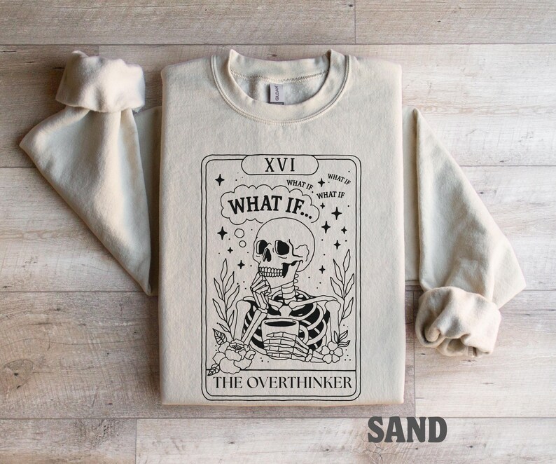 Puede incluir: Sudadera color arena con un dise&ntilde;o de carta de tarot en blanco y negro. La carta representa un esqueleto sosteniendo una taza, con el texto "WHAT IF..." y "THE OVERTHINKER". Las mangas est&aacute;n dobladas.