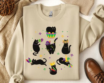 May include: A tan sweatshirt with a black cat design. The cats are wearing Mardi Gras masks, hats, and holding Mardi Gras beads and other festive items. The design is surrounded by green, purple, and gold Mardi Gras colors.