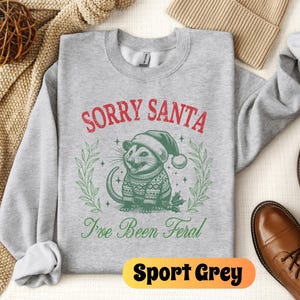 May include: A gray sweatshirt with the text "Sorry Santa I've Been Feral" in red and green. A cartoon possum wearing a Santa hat is in the center of the text. The sweatshirt is on a white background.