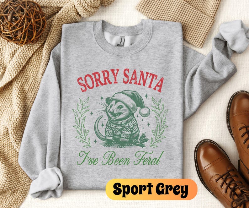 May include: A gray sweatshirt with the text "Sorry Santa I've Been Feral" in red and green. A cartoon possum wearing a Santa hat is in the center of the text. The sweatshirt is on a white background.