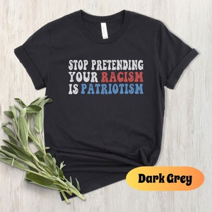 May include: Black t-shirt with the text "STOP PRETENDING YOUR RACISM IS PATRIOTISM" in red, white, and blue lettering. The shirt is on a light wood background.