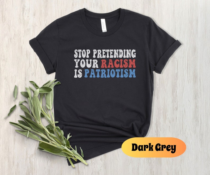 May include: Black t-shirt with the text "STOP PRETENDING YOUR RACISM IS PATRIOTISM" in red, white, and blue lettering. The shirt is on a light wood background.