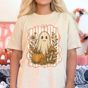 May include: Beige t-shirt featuring a Halloween-themed graphic. The design includes a smiling ghost, pumpkin, mushrooms, and fall foliage within a striped frame. The shirt is a neutral color, suitable for autumn.