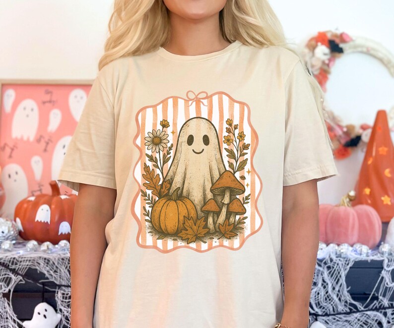 May include: Beige t-shirt featuring a Halloween-themed graphic. The design includes a smiling ghost, pumpkin, mushrooms, and fall foliage within a striped frame. The shirt is a neutral color, suitable for autumn.