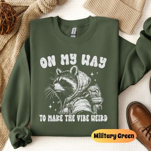 May include: A green sweatshirt with a white raccoon graphic and the text "On My Way To Make The Vibe Weird". The sweatshirt is a crew neck style and has a relaxed fit. The color of the sweatshirt is Military Green.