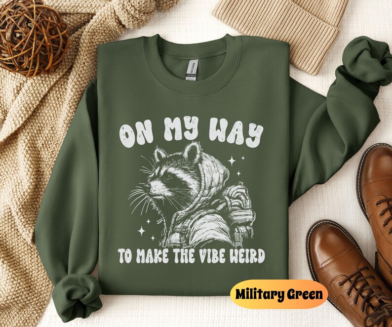 May include: A green sweatshirt with a white raccoon graphic and the text "On My Way To Make The Vibe Weird". The sweatshirt is a crew neck style and has a relaxed fit. The color of the sweatshirt is Military Green.