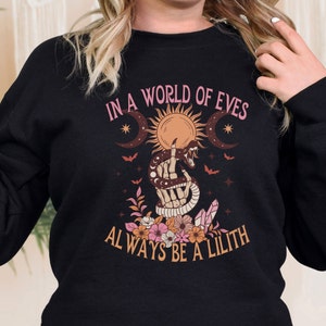 May include: Black sweatshirt with a graphic design featuring a snake wrapped around a hand with a moon, sun, and stars. The text reads "In a world of Eves, always be a Lilith."