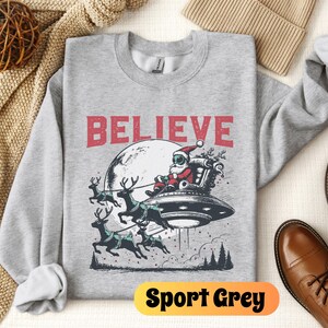 May include: A gray sweatshirt with a Christmas-themed graphic featuring Santa Claus in a spaceship with reindeer pulling the ship. The text "BELIEVE" is printed above the graphic. The sweatshirt is labeled "Sport Grey".