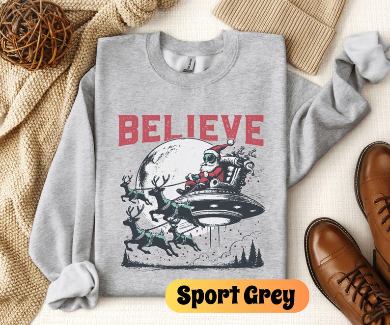 May include: A gray sweatshirt with a Christmas-themed graphic featuring Santa Claus in a spaceship with reindeer pulling the ship. The text "BELIEVE" is printed above the graphic. The sweatshirt is labeled "Sport Grey".