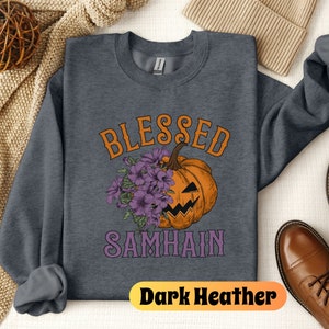 以下が含まれることがあります： 濃いグレーのヘザーのトレーナーに、紫とオレンジ色の「Blessed Samhain」という文字が書かれています。文字の中央には、紫の花がついたジャック・オー・ランタンがあります。