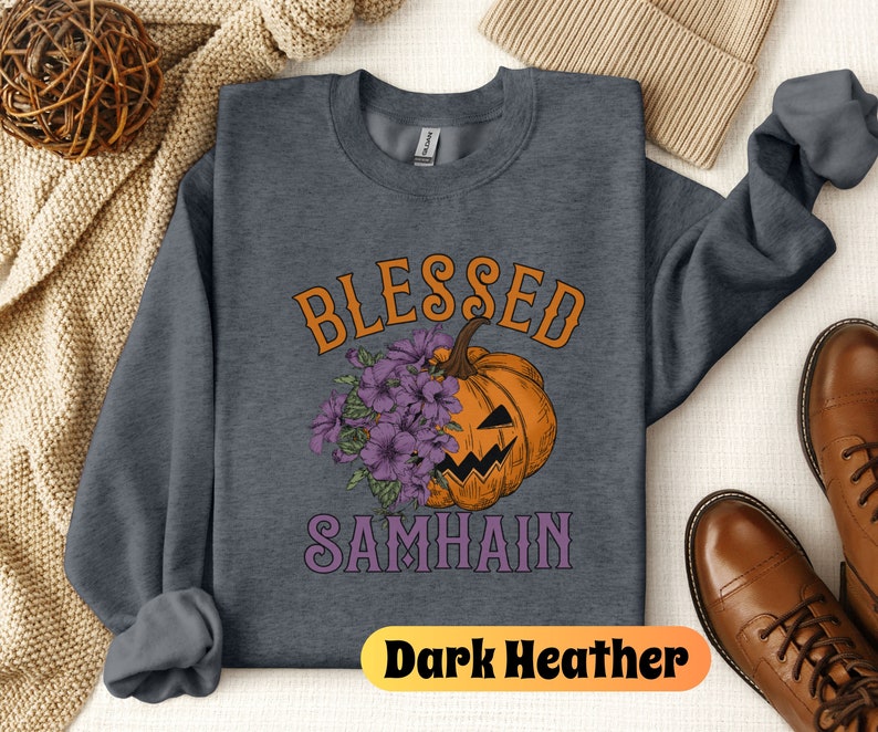 以下が含まれることがあります： 濃いグレーのヘザーのトレーナーに、紫とオレンジ色の「Blessed Samhain」という文字が書かれています。文字の中央には、紫の花がついたジャック・オー・ランタンがあります。