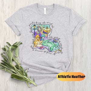 Op de afbeelding: Een grijs T-shirt met een kleurrijke illustratie van de staat Louisiana. De illustratie bevat verschillende symbolen en iconen die geassocieerd worden met Mardi Gras, zoals een koningentaart, een fleur-de-lis en een paradewagen. De tekst "Mardi Gras" is ook opgenomen in de illustratie. Het T-shirt is gemarkeerd met "Athletic Heather".