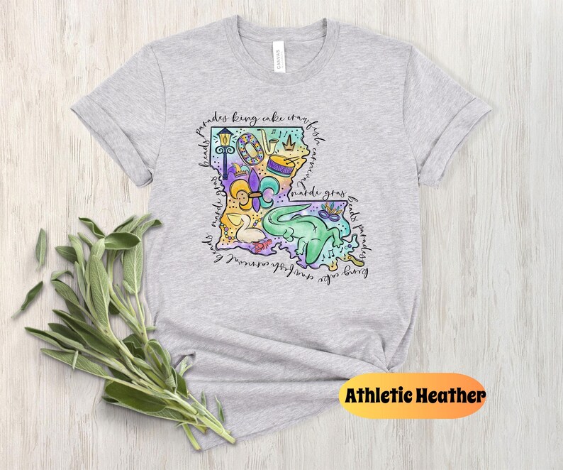 Op de afbeelding: Een grijs T-shirt met een kleurrijke illustratie van de staat Louisiana. De illustratie bevat verschillende symbolen en iconen die geassocieerd worden met Mardi Gras, zoals een koningentaart, een fleur-de-lis en een paradewagen. De tekst "Mardi Gras" is ook opgenomen in de illustratie. Het T-shirt is gemarkeerd met "Athletic Heather".
