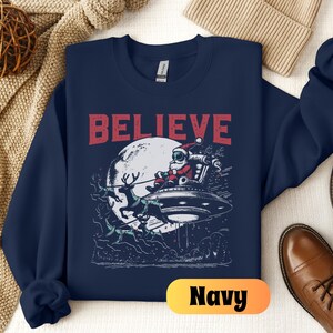 May include: Navy blue sweatshirt with a graphic of Santa Claus flying in a UFO with reindeer. The text "BELIEVE" is printed in red above the graphic.