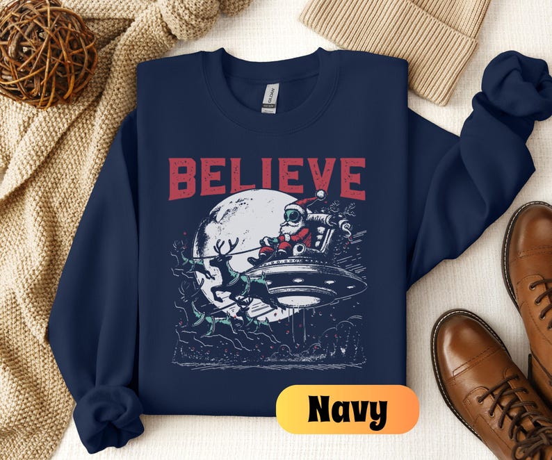 May include: Navy blue sweatshirt with a graphic of Santa Claus flying in a UFO with reindeer. The text "BELIEVE" is printed in red above the graphic.