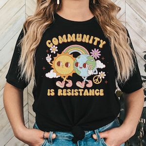 May include: Black t-shirt with a graphic of a sun and earth holding hands with a rainbow and the text "Community is Resistance".