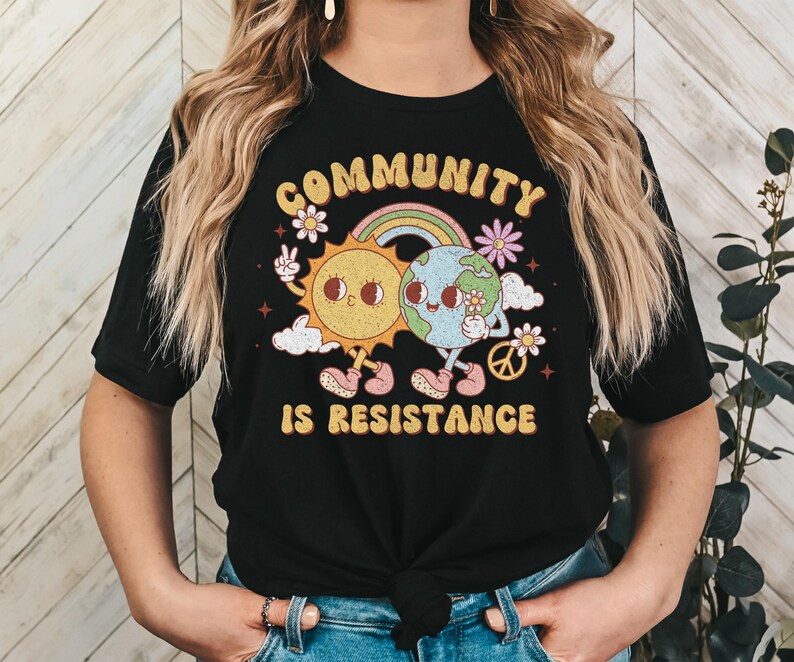 May include: Black t-shirt with a graphic of a sun and earth holding hands with a rainbow and the text "Community is Resistance".