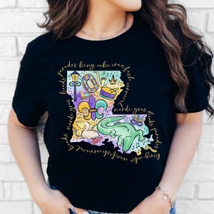 Op de afbeelding: Zwart T-shirt met een kleurrijke illustratie van de staat Louisiana. De illustratie bevat Mardi Gras-parels, een koningentaart, een fleur-de-lis, een trompet, een trommel, een masker en een alligator. De tekst "beads parades king cake crawfish carnival mardi gras beads parades" omringt de illustratie.