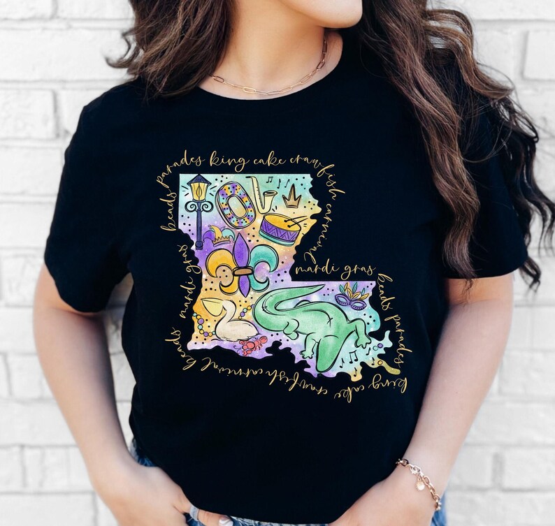 Op de afbeelding: Zwart T-shirt met een kleurrijke illustratie van de staat Louisiana. De illustratie bevat Mardi Gras-parels, een koningentaart, een fleur-de-lis, een trompet, een trommel, een masker en een alligator. De tekst "beads parades king cake crawfish carnival mardi gras beads parades" omringt de illustratie.