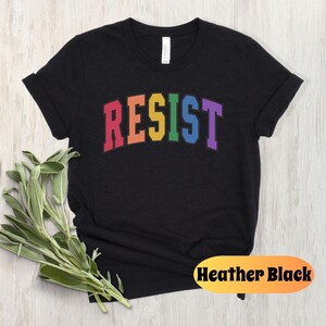 May include: Black t-shirt with the word "RESIST" in rainbow colors. The word is in a collegiate font style.