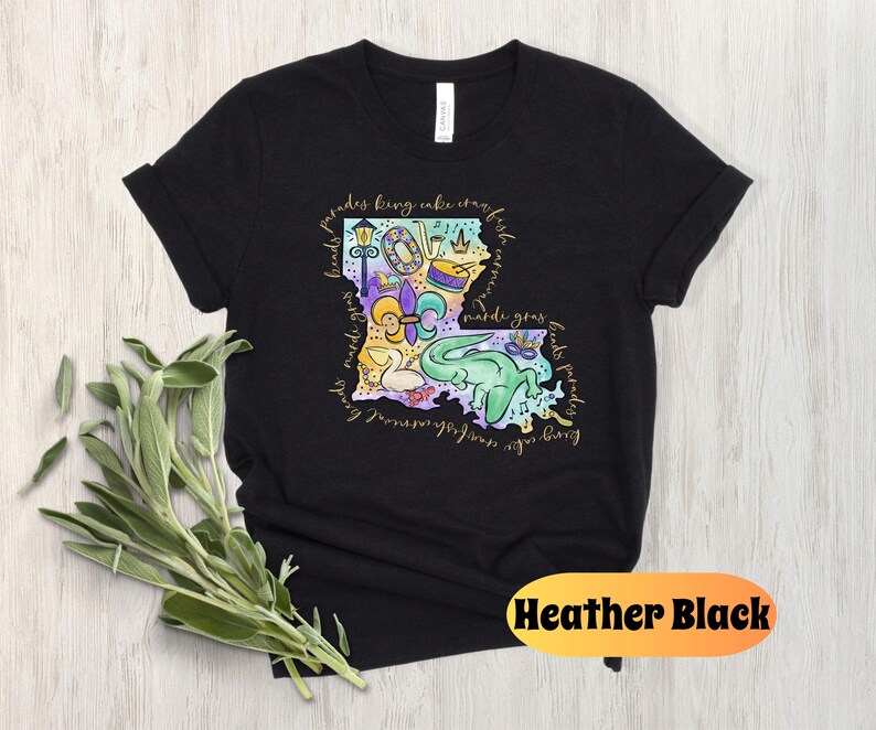 Op de afbeelding: Zwart T-shirt met een kleurrijke illustratie van de staat Louisiana. De illustratie bevat Mardi Gras-parels, een koningentaart, een fleur-de-lis, een trommel, een paradewagen en een alligator. De tekst "parades king cake crawfish first friday mardi gras floats parades" staat rond de omtrek van de staat.