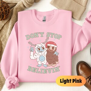 May include: Light pink sweatshirt with a graphic of a yeti and a brown monster with the text "Don't Stop Believin'" printed on the front.