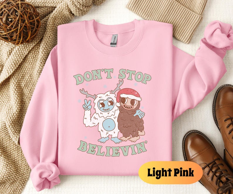 May include: Light pink sweatshirt with a graphic of a yeti and a brown monster with the text "Don't Stop Believin'" printed on the front.