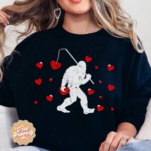 May include: Black sweatshirt with a white graphic of a Bigfoot holding a heart-shaped fishing lure. The graphic is surrounded by red hearts.