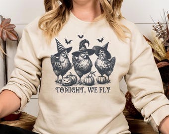 May include: A cream-colored sweatshirt with a graphic of three chickens wearing witch hats and holding pumpkins. The text "Tonight, We Fly" is printed below the graphic.