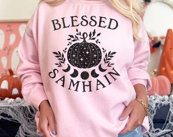 以下が含まれることがあります： 黒い文字で「Blessed Samhain」と書かれたピンクのトレーナー。デザインには、星と三日月が描かれた黒いカボチャが描かれています。