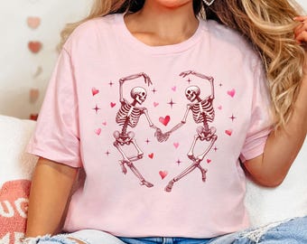 May include: Pink t-shirt with a graphic of two skeletons dancing with hearts and stars. The skeletons are holding hands and smiling.