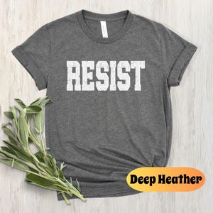 May include: Deep heather grey t-shirt featuring the word "RESIST" in white block letters. The shirt has a crew neck and short sleeves. A sprig of green leaves is placed on the left side of the shirt. The words "Deep Heather" are printed on an orange oval.