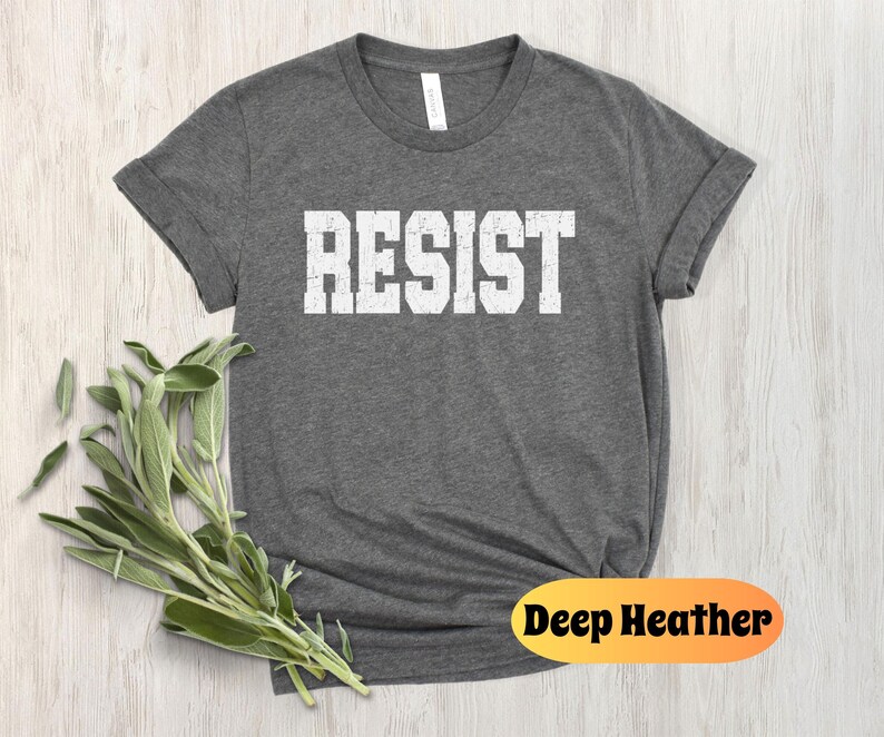 May include: Deep heather grey t-shirt featuring the word "RESIST" in white block letters. The shirt has a crew neck and short sleeves. A sprig of green leaves is placed on the left side of the shirt. The words "Deep Heather" are printed on an orange oval.