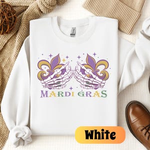May include: White sweatshirt with a purple and green Mardi Gras design featuring two skeletal hands holding fleur-de-lis. The text "Mardi Gras" is printed in green and yellow.